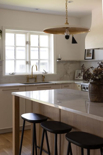 Cuisine haut de gamme conçue par Ateliers Jacob à Montréal, mariant des armoires crème avec des poignées en laiton, un îlot en bois clair et un comptoir en marbre. La hotte sculpturale et les luminaires suspendus ajoutent une touche artisanale et contemporaine à l’ensemble.