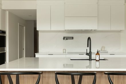 Cuisine moderne Ateliers Jacob avec comptoir en quartz blanc, grand îlot central en bois clair avec chaises noires, armoires pleine hauteur intégrées et robinetterie noire mate.