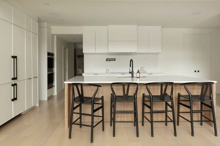 Cuisine moderne Ateliers Jacob avec comptoir en quartz blanc, grand îlot central en bois clair avec chaises noires, armoires pleine hauteur intégrées et robinetterie noire mate.