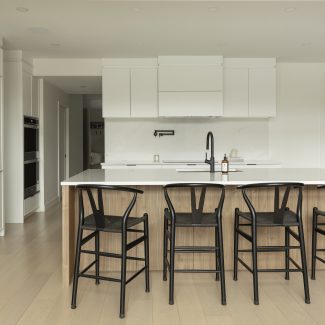 uisine moderne Ateliers Jacob avec comptoir en quartz blanc, grand îlot central en bois clair avec chaises noires, armoires pleine hauteur intégrées et robinetterie noire mate.