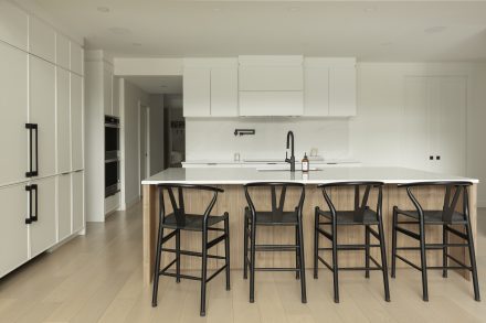 uisine moderne Ateliers Jacob avec comptoir en quartz blanc, grand îlot central en bois clair avec chaises noires, armoires pleine hauteur intégrées et robinetterie noire mate.