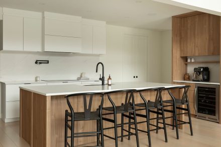 Cuisine moderne Ateliers Jacob avec comptoir en quartz blanc, grand îlot central en bois clair avec chaises noires, armoires pleine hauteur intégrées et robinetterie noire mate.