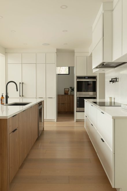 Cuisine moderne Ateliers Jacob avec comptoir en quartz blanc, grand îlot central en bois clair avec chaises noires, armoires pleine hauteur intégrées et robinetterie noire mate.