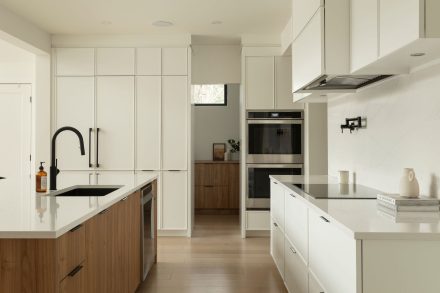 Cuisine moderne Ateliers Jacob avec comptoir en quartz blanc, grand îlot central en bois clair avec chaises noires, armoires pleine hauteur intégrées et robinetterie noire mate.