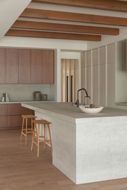 Cuisine moderne haut de gamme signée Ateliers Jacob avec îlot en pierre claire, hotte dissimulée en béton, armoires pleine hauteur crème, placages de bois naturel et poutres apparentes au plafond, design minimaliste et chaleureux.