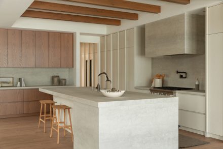 Cuisine moderne haut de gamme signée Ateliers Jacob avec îlot en pierre claire, hotte dissimulée en béton, armoires pleine hauteur crème, placages de bois naturel et poutres apparentes au plafond, design minimaliste et chaleureux.
