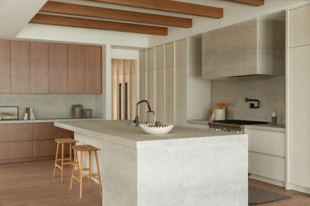 Cuisine moderne haut de gamme signée Ateliers Jacob avec îlot en pierre claire, hotte dissimulée en béton, armoires pleine hauteur crème, placages de bois naturel et poutres apparentes au plafond, design minimaliste et chaleureux.