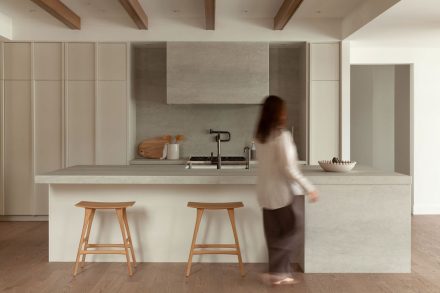 Cuisine moderne haut de gamme signée Ateliers Jacob avec îlot en pierre claire, hotte dissimulée en béton, armoires pleine hauteur crème, placages de bois naturel et poutres apparentes au plafond, design minimaliste et chaleureux.