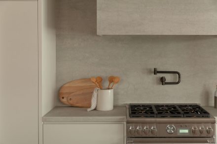 Cuisine moderne haut de gamme signée Ateliers Jacob avec îlot en pierre claire, hotte dissimulée en béton, armoires pleine hauteur crème, placages de bois naturel et poutres apparentes au plafond, design minimaliste et chaleureux.