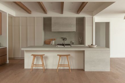 Cuisine moderne haut de gamme signée Ateliers Jacob avec îlot en pierre claire, hotte dissimulée en béton, armoires pleine hauteur crème, placages de bois naturel et poutres apparentes au plafond, design minimaliste et chaleureux.