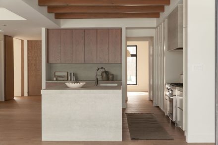 Cuisine moderne haut de gamme signée Ateliers Jacob avec îlot en pierre claire, hotte dissimulée en béton, armoires pleine hauteur crème, placages de bois naturel et poutres apparentes au plafond, design minimaliste et chaleureux.
