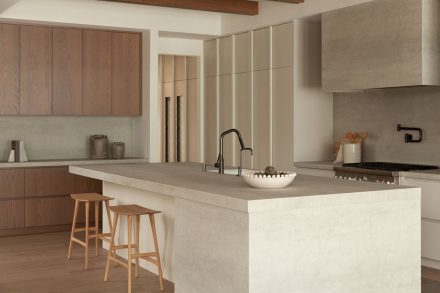 Cuisine moderne haut de gamme signée Ateliers Jacob avec îlot en pierre claire, hotte dissimulée en béton, armoires pleine hauteur crème, placages de bois naturel et poutres apparentes au plafond, design minimaliste et chaleureux.