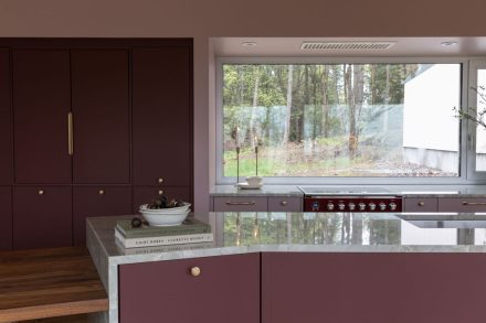 Cuisine Ateliers Jacob avec armoires bourgogne sur mesure et comptoir en pierre naturelle, îlot central et grande fenêtre ouverte sur la forêt