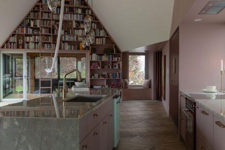 Cuisine sur mesure Ateliers Jacob avec îlot en pierre naturelle, armoires bourgogne mates, robinetterie en laiton et bibliothèque intégrée dans un espace lumineux