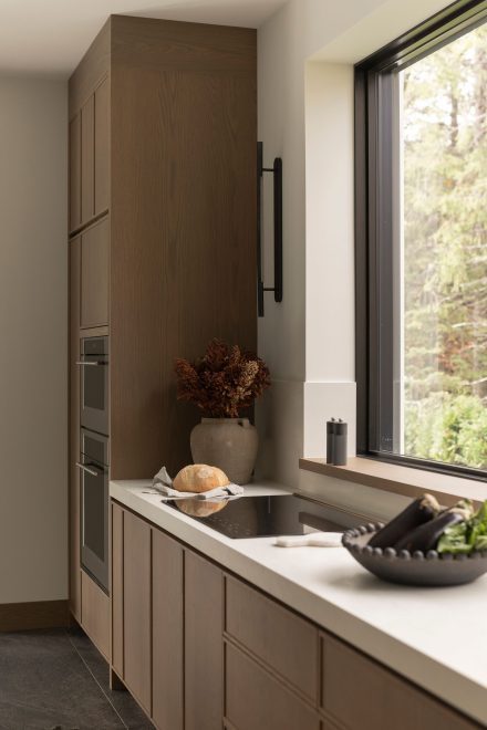 Cuisine sur mesure en chêne conçue par Ateliers Jacob, où la lumière naturelle entre par une large fenêtre et éclaire les surfaces en bois et en pierre, avec vue sur les arbres.