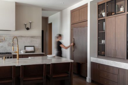 Cuisine sur mesure avec îlot en pierre naturelle, armoires en bois foncé, vitrines encadrées de verre et réfrigérateur intégré dissimulé derrière des panneaux assortis.