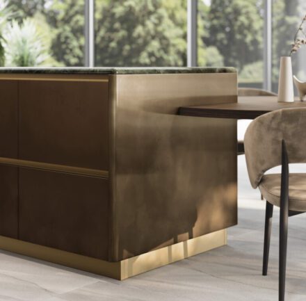 Îlot de cuisine aux courbes adoucies avec table intégrée, façades métalliques brunies et plateau en marbre Verde Borgogna aux veines profondes, lumière naturelle révélant la matière. Projet: GF Interiors par Guzzini & Fontana.