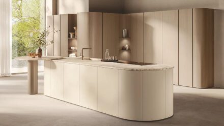 Cuisine Scavolini présentée à l’EuroCucina 2026, îlot aux lignes arrondies en finition crème et plan de travail en pierre claire, lumière naturelle et bois clair en arrière-plan