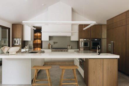 Cuisine contemporaine haut de gamme aux lignes épurées, combinant armoires en bois naturel et surfaces blanches, avec îlot central fonctionnel, hotte architecturale encastrée et électroménagers intégrés, créant un espace lumineux et équilibré pensé pour l’ergonomie et la fluidité des usages.