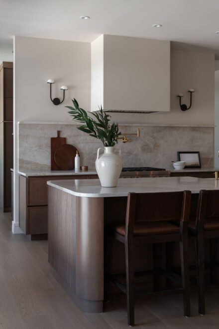 Cuisine Ateliers Jacob avec îlot arrondi en quartzite naturel, armoires en bois foncé sur mesure et hotte minimaliste dans une cuisine haut de gamme chaleureuse