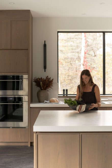 Cuisine haut de gamme Ateliers Jacob avec îlot en porcelaine, armoires en chêne et grande fenêtre ouverte sur la forêt, mettant en scène un moment du quotidien dans un espace lumineux et épuré.