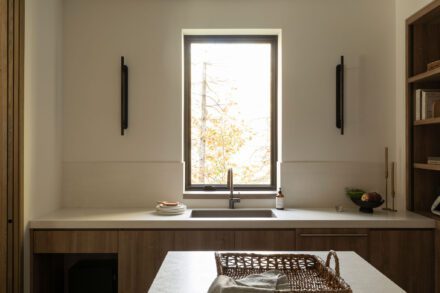 Cuisine-arrière Ateliers Jacob en bois de chêne avec comptoir de porcelaine, évier intégré sous une fenêtre pleine hauteur ouverte sur la forêt de Saint-Sauveur, réalisée avec Symbiose Design.