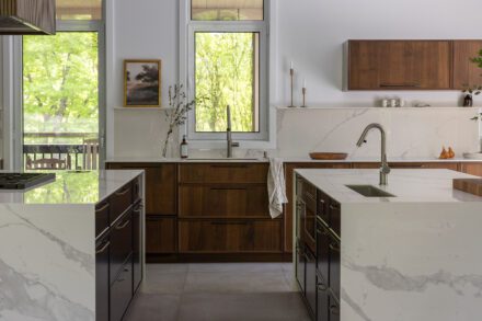 Cuisine haut de gamme Ateliers Jacob avec deux îlots en Dekton effet marbre, armoires en noyer foncé et grande fenestration ouverte sur la nature.