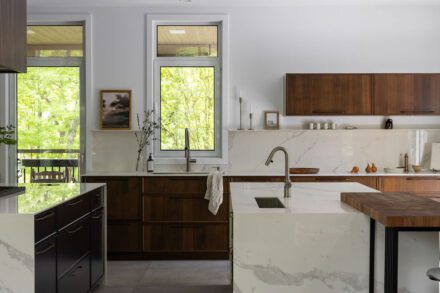 Cuisine haut de gamme Ateliers Jacob avec deux îlots en Dekton effet marbre, armoires en noyer foncé et grande fenestration ouverte sur la nature.