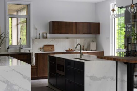 Cuisine haut de gamme Ateliers Jacob avec deux îlots en Dekton effet marbre, armoires en noyer foncé et grande fenestration ouverte sur la nature.