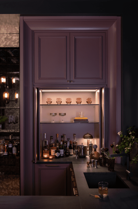 Custom speakeasy bar by Ateliers Jacob: plum cabinetry, backlit glass shelves and brass details create an intimate, elegant cocktail nook.