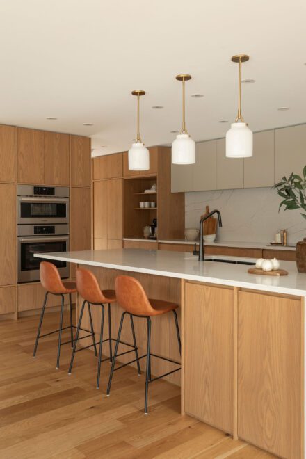 Cuisine Ateliers Jacob en chêne avec grand îlot et comptoir de quartz blanc, évier noir, fours encastrés, luminaires suspendus et tabourets en cuir cognac.